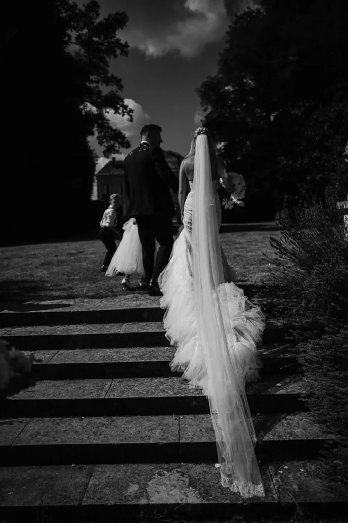 A bride and her family walk up stone steps. Her long veils trails behind her.