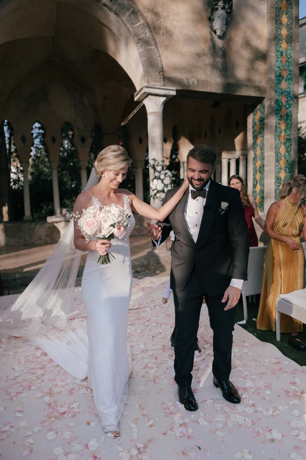 Melanie and Ruben walking down a rose petal-strewn aisle, smiling joyfully at their Villa Cimbrone wedding, with historic arches and columns in the background.