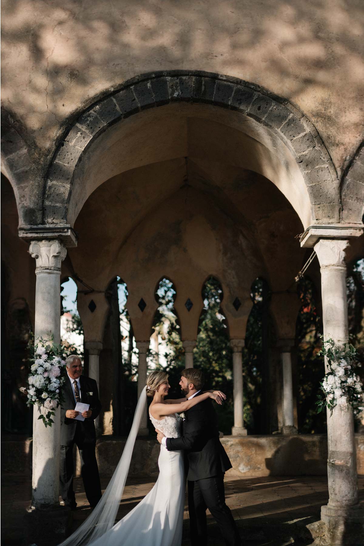 Melanie and Ruben share a tender embrace under the ancient arches during their Villa Cimbrone wedding, with soft light filtering through gothic windows and the officiant smiling warmly