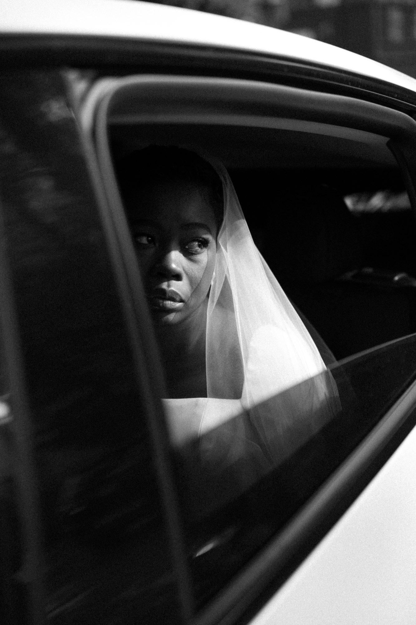 A bride wearing a veil looks thoughtfully out of a car window at her Kensington Temple wedding. The image is in black and white, capturing an intimate and contemplative moment.