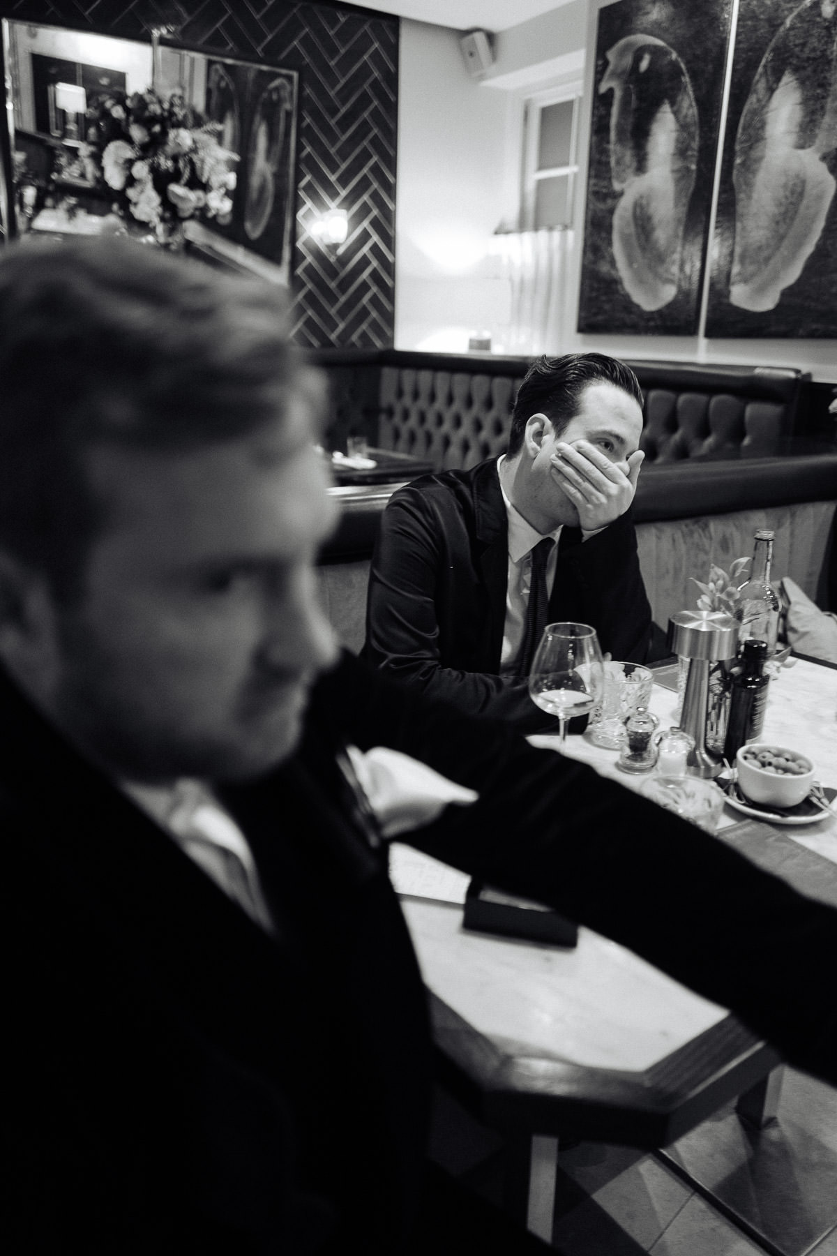 Best man with hand to mouth sitting behind the groom in a Mayfair restaurant on the morning of the wedding, capturing a candid moment.