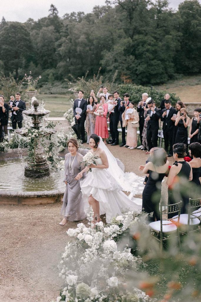 A bride and her mother walk past a fountain as they head towards their outdoor ceremony, taken by London Wedding Photographer Philip White.