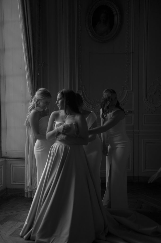 A bride looks out of the window as three of her bridesmaids help her into her wedding dress, taken by London Wedding Photographer Philip White.