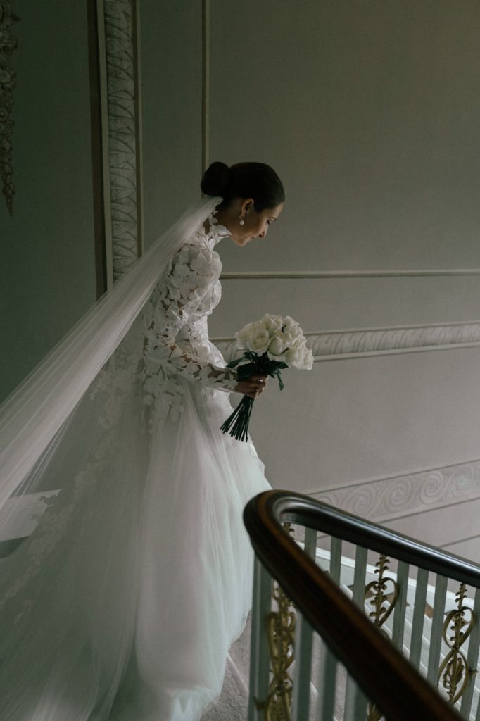 a bride, holding her bouquet begins to descend the stair case of an English stately home, taken by London Wedding Photographer Philip White.